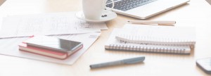 Collection of work materials including sheets of paper, several notebooks, a phone, pens, a cup of coffee, and a laptop