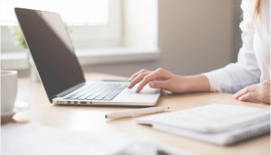 Close up of a woman using the touchpad on her laptop with a notebook, pen, and cup of coffee