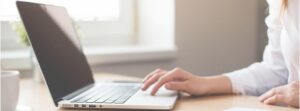 Close up of a woman using the touchpad on her laptop with a notebook, pen, and cup of coffee